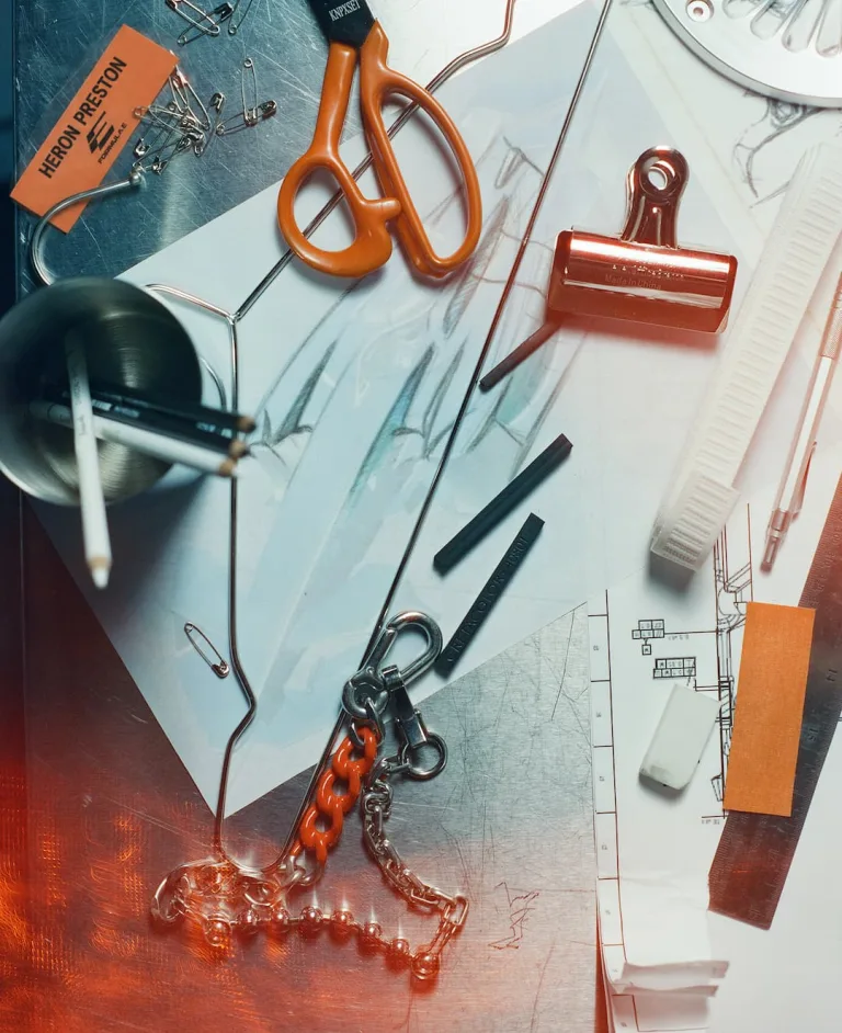 Heron Preston Formula E partnership flat-lay showing orange scissors, a copper bulldog clip, safety pins, chains, pencils and a Heron Preston Formula E tag laid over race car sketches and technical drawings on a design desk ©Formula E